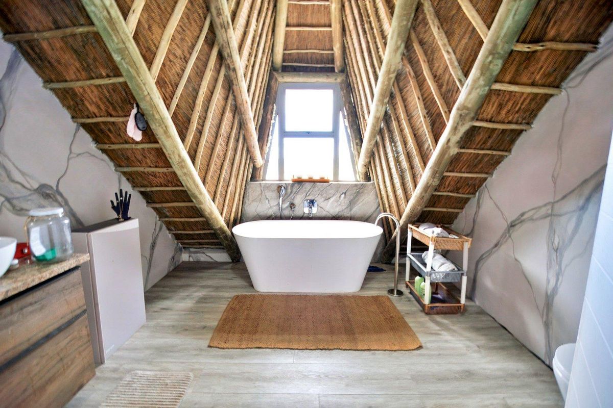 A unique bathroom featuring a freestanding bathtub, exposed timber rafters, thatched roof detailing, and modern tiled flooring under a pitched ceiling.