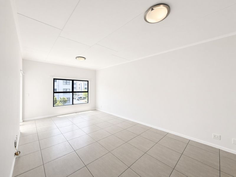 A bright, empty living area featuring neutral tiled flooring, clean white walls, and a large window providing natural light.