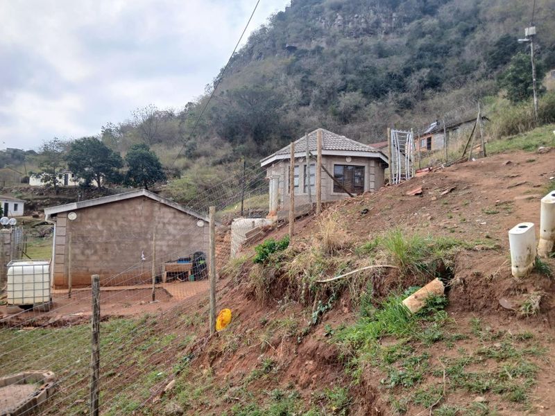 Exterior view of a residential property featuring concrete block structures, tiled roofing, and wire fencing set against a sloped, undeveloped hillside.
