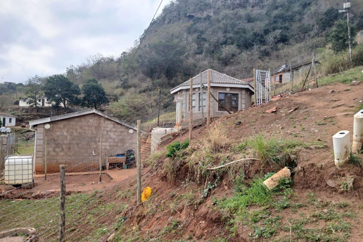 Exterior view of a residential property featuring concrete block structures, tiled roofing, and wire fencing set against a sloped, undeveloped hillside.