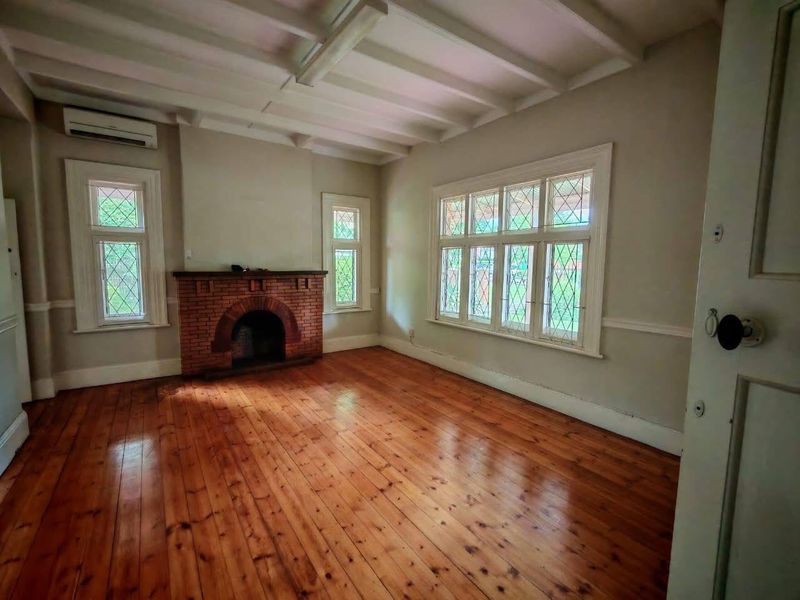 A bright, empty office space featuring original timber flooring, a classic brick fireplace, exposed ceiling beams, and charming leaded glass windows.