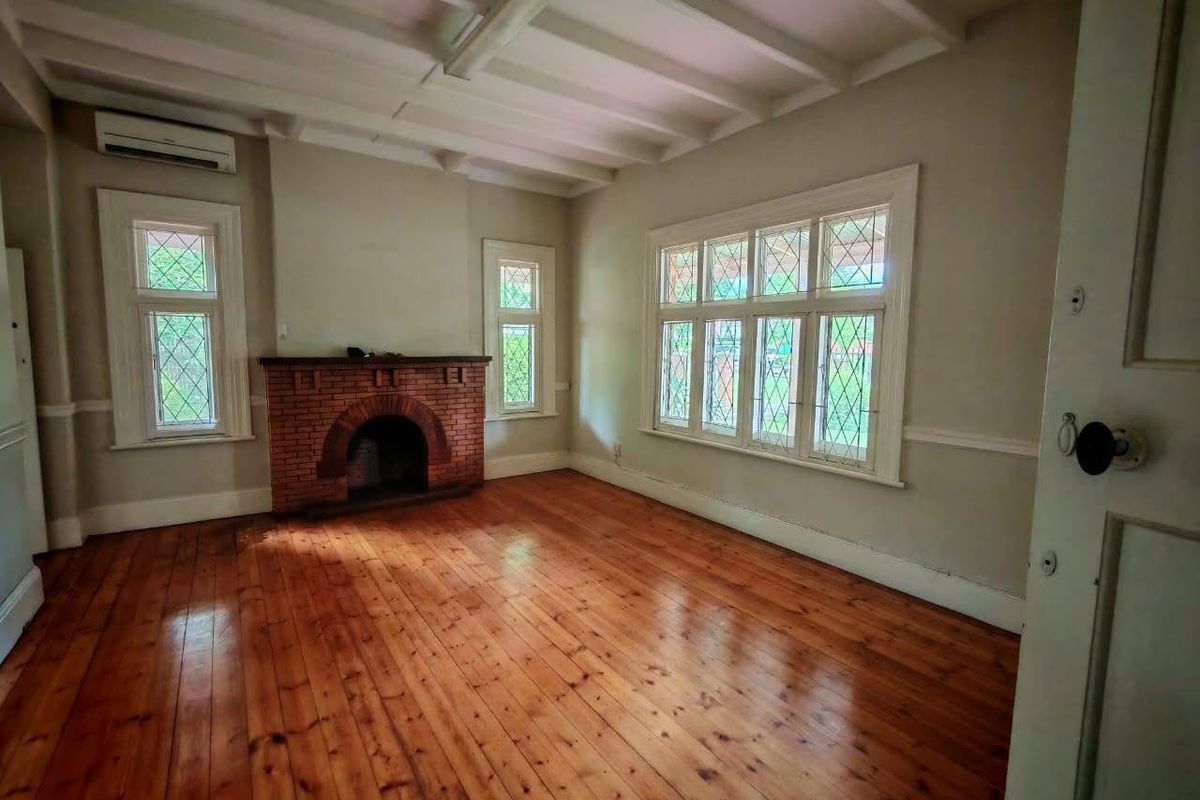 A bright, empty office space featuring original timber flooring, a classic brick fireplace, exposed ceiling beams, and charming leaded glass windows.