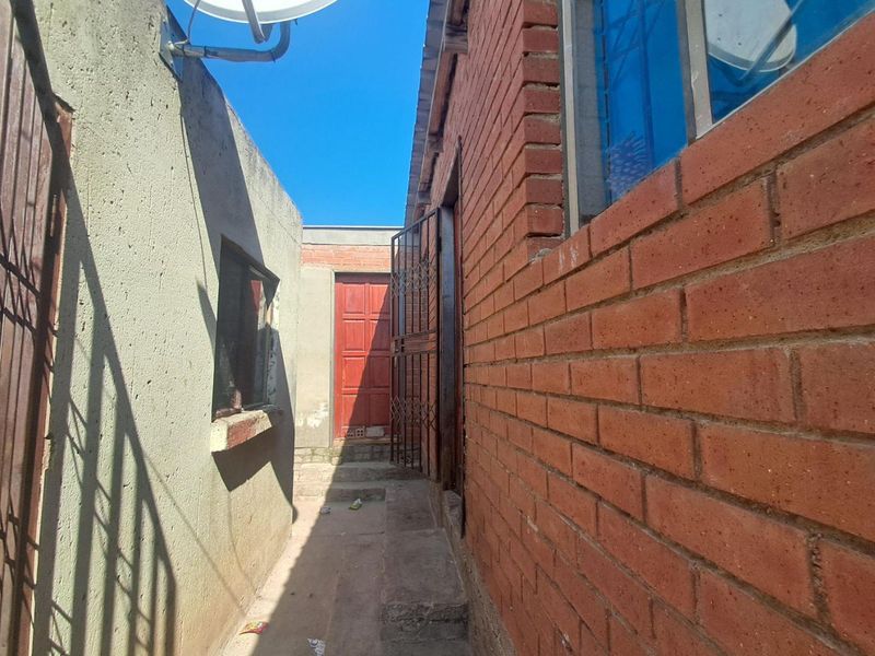 A narrow exterior side passage featuring exposed brick walls, a security gate, and a concrete walkway leading to a rear entrance.