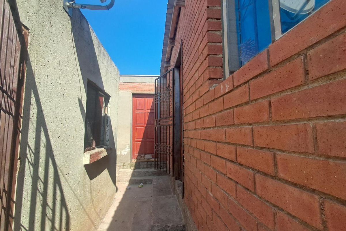 A narrow exterior side passage featuring exposed brick walls, a security gate, and a concrete walkway leading to a rear entrance.