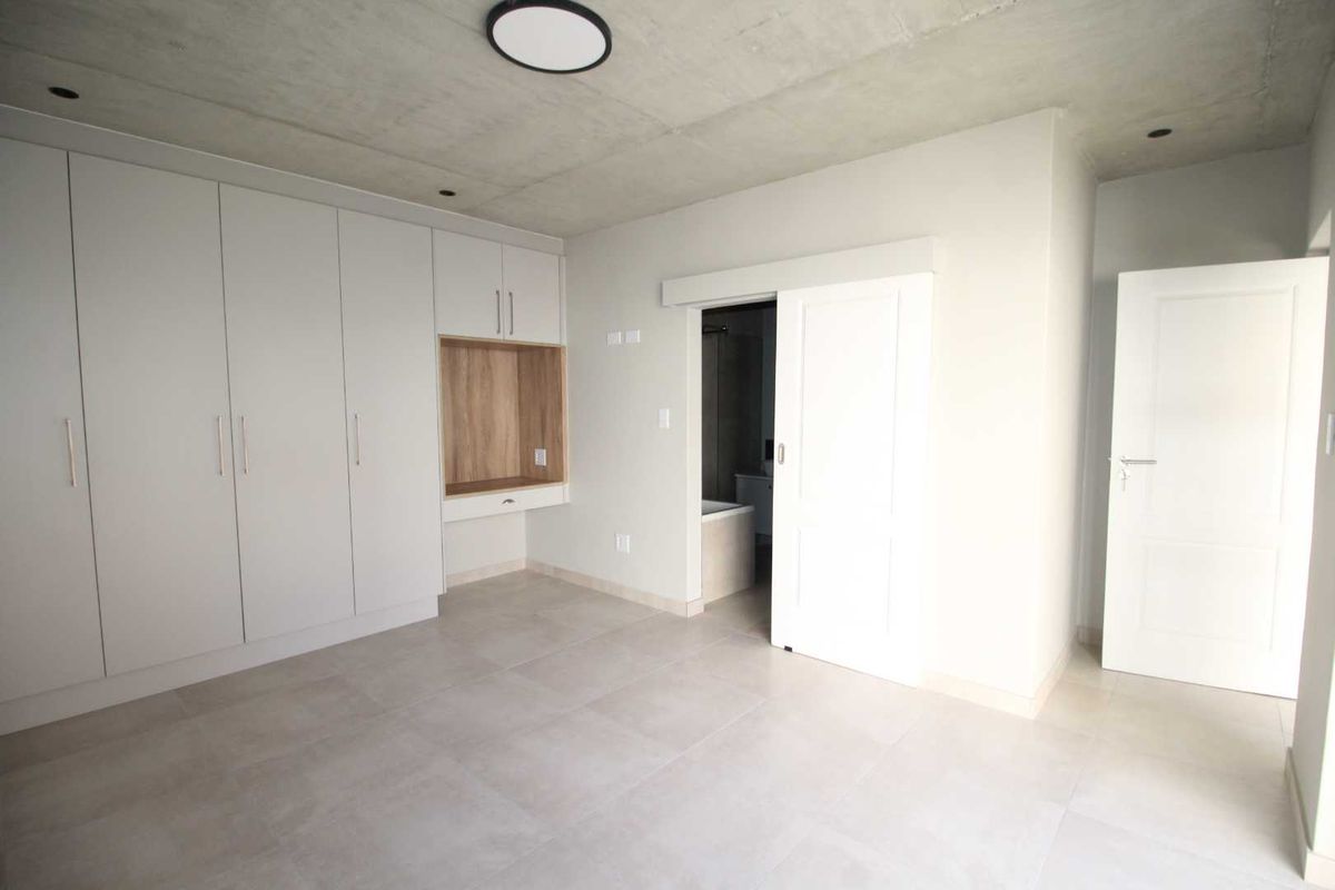 Modern bedroom featuring built-in wardrobes with integrated desk space, exposed concrete ceiling, tiled flooring, and an en-suite bathroom access via a sliding door.