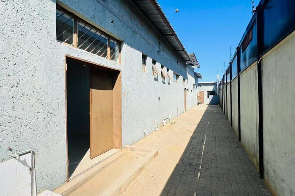 A narrow paved exterior side passage featuring a concrete wall, electric fencing, and basic drainage infrastructure along the building exterior.