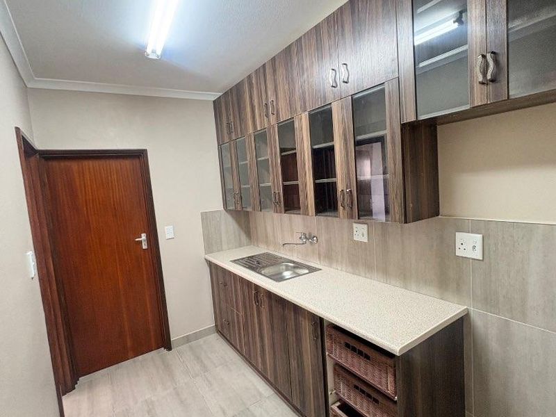 A clean and functional scullery area featuring modern wood-grain cabinetry, stone-look countertops, a stainless steel sink, and neutral wall tiling.