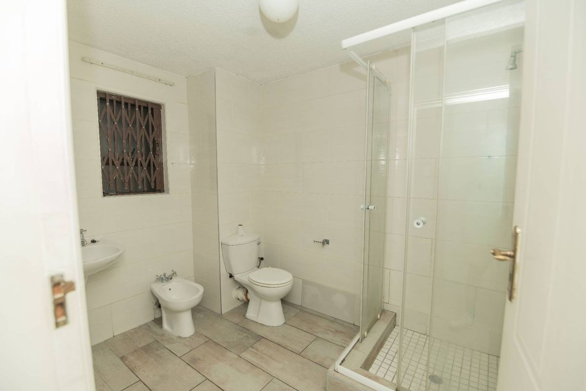 A functional bathroom featuring a glass-enclosed shower, toilet, bidet, and wall-mounted basin with neutral wall tiling and wood-look floor tiles.