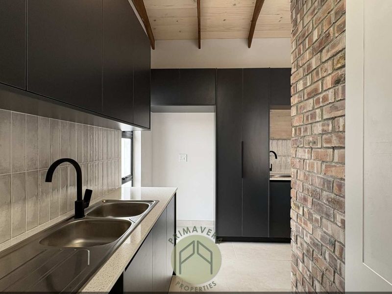Modern scullery featuring sleek black cabinetry, stone countertops, double sink, and an exposed brick feature wall with timber ceiling beams.