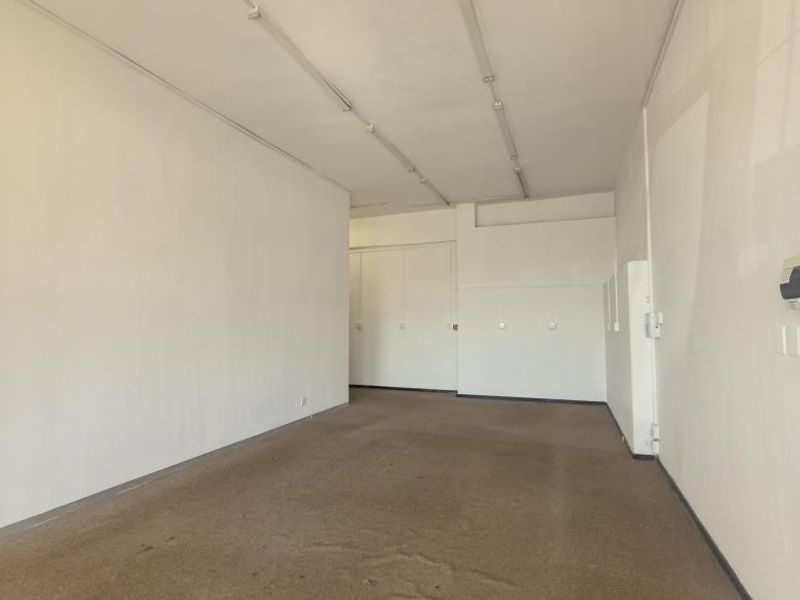 An empty, open-plan office space featuring neutral walls, fluorescent strip lighting, and built-in storage cupboards with worn carpeting throughout.