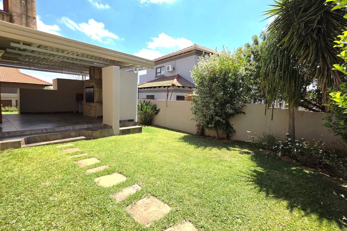 A well-maintained rear garden featuring a covered patio area with a built-in braai, stone stepping stones, and established perimeter walling.