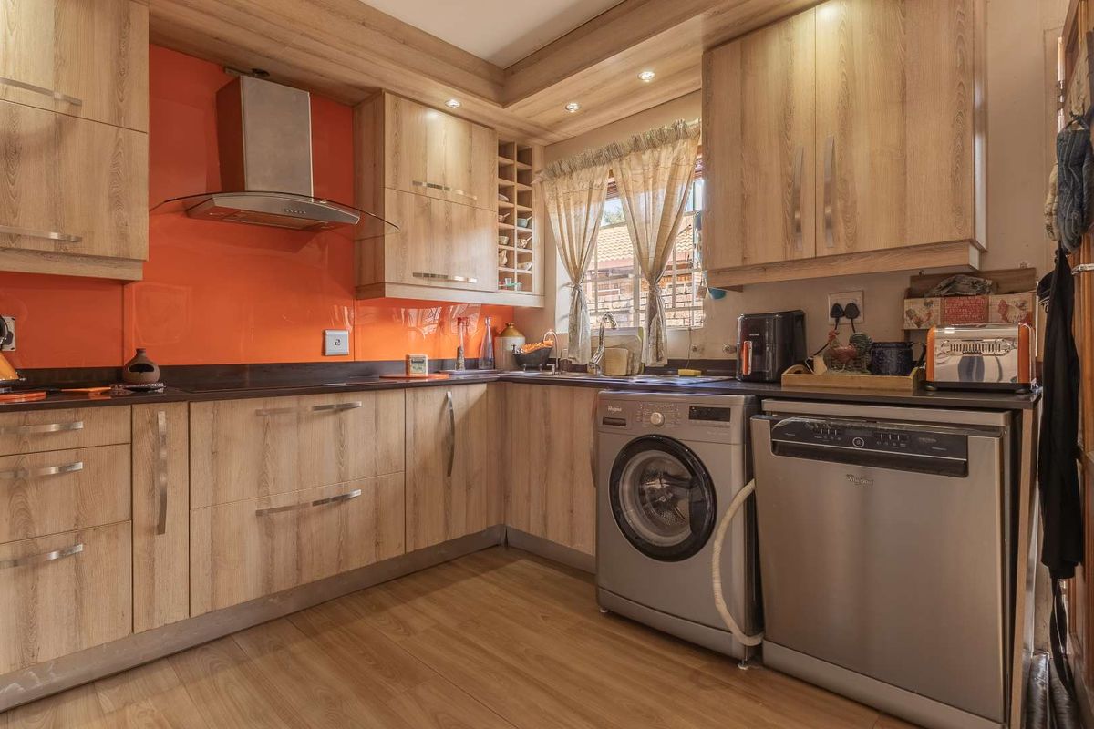 A functional kitchen featuring wood-look cabinetry, a vibrant orange glass splashback, integrated extractor fan, and dedicated spaces for laundry and dishwasher appliances.