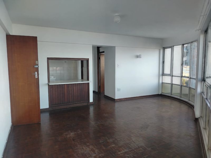 An empty living area featuring original parquet flooring, a timber serving hatch to the kitchen, and large windows providing natural light.