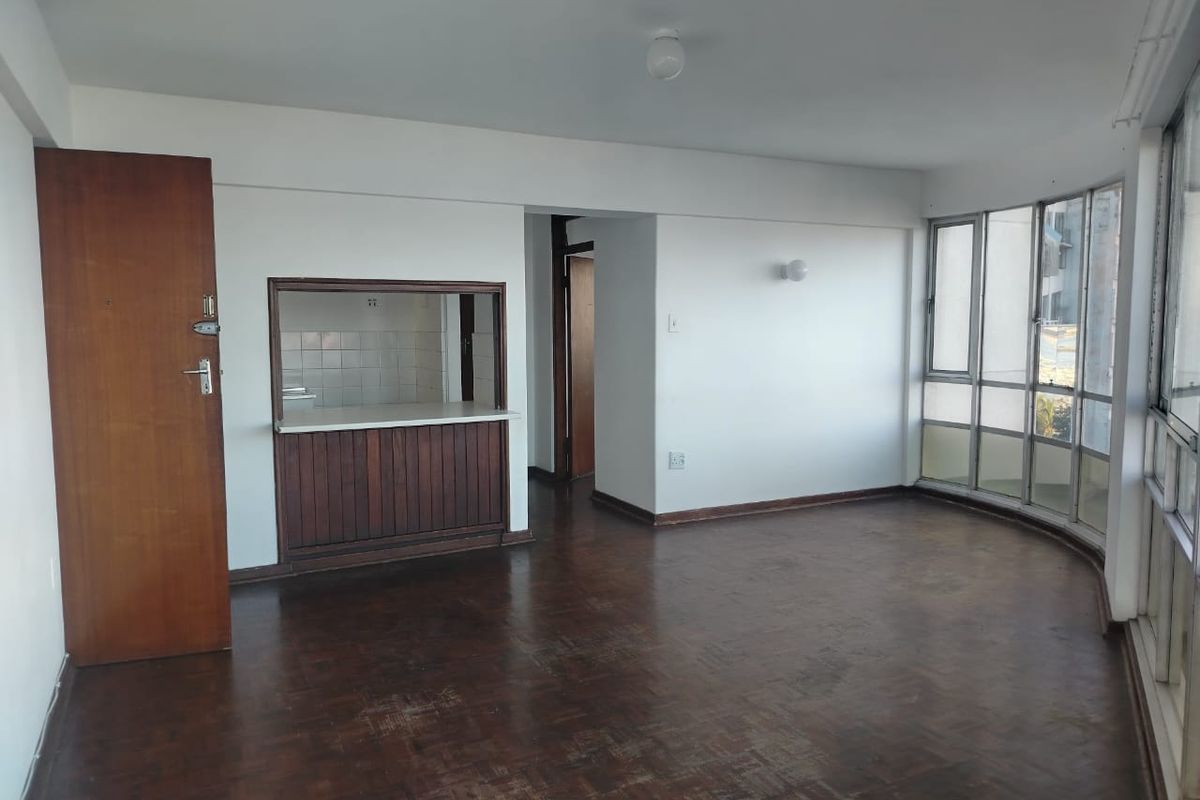 An empty living area featuring original parquet flooring, a timber serving hatch to the kitchen, and large windows providing natural light.
