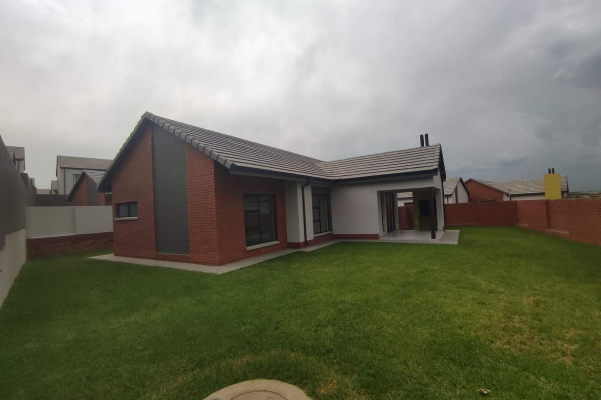 Rear view of a modern brick house featuring a covered patio, tiled roof, and a well-maintained lawn within a secure boundary wall.