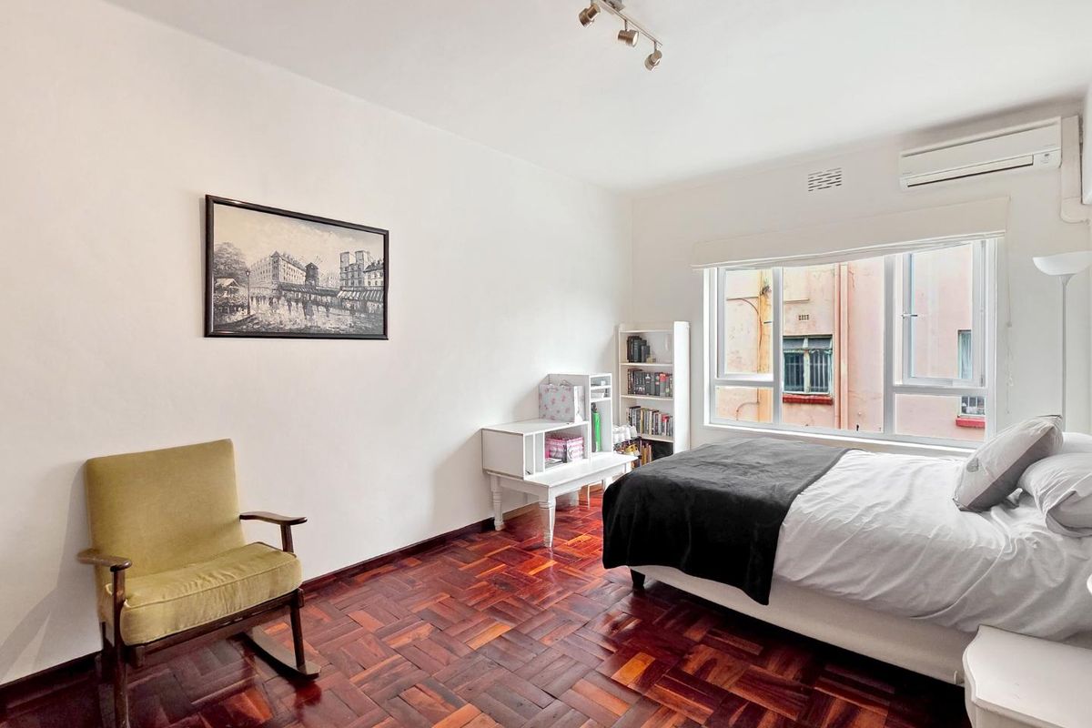 A well-lit bedroom featuring polished parquet flooring, a wall-mounted split-system air conditioner, and large windows providing natural light.