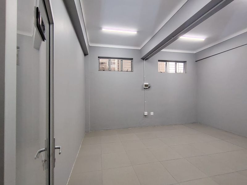 A clean, empty office space featuring neutral grey walls, tiled flooring, exposed structural beams, and functional fluorescent lighting.
