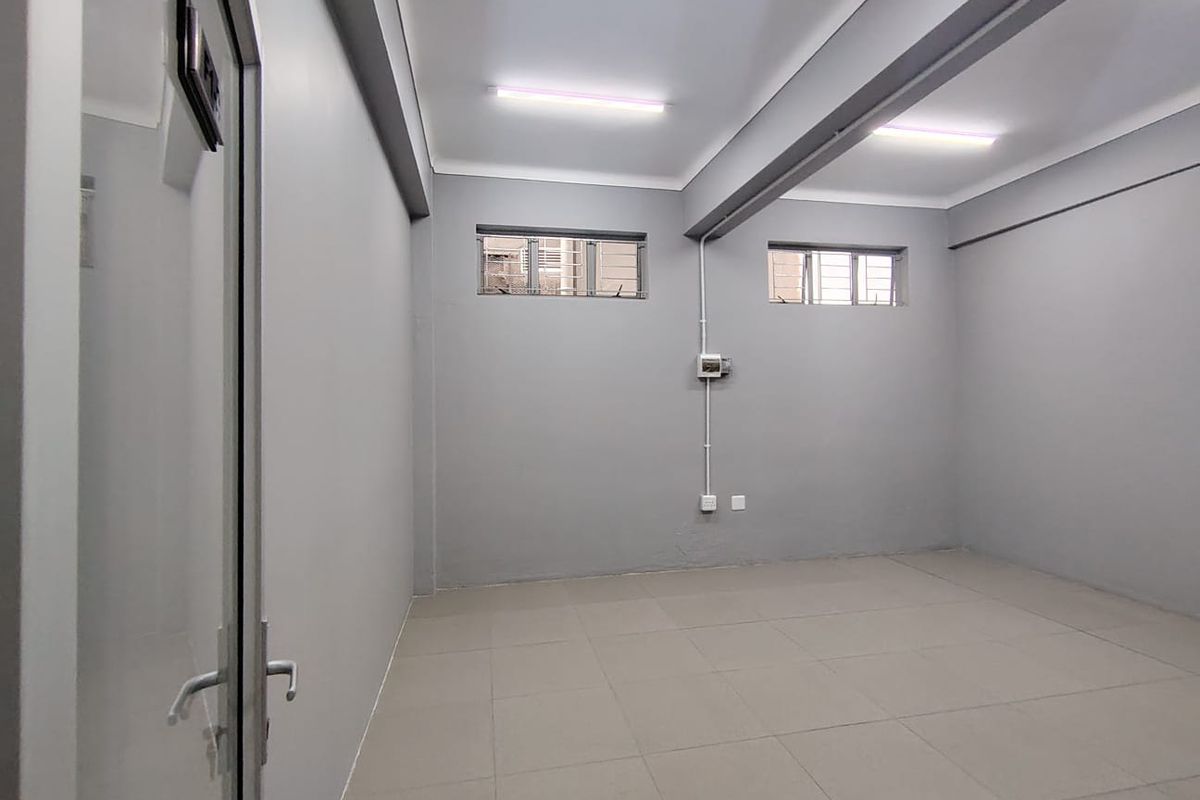 A clean, empty office space featuring neutral grey walls, tiled flooring, exposed structural beams, and functional fluorescent lighting.