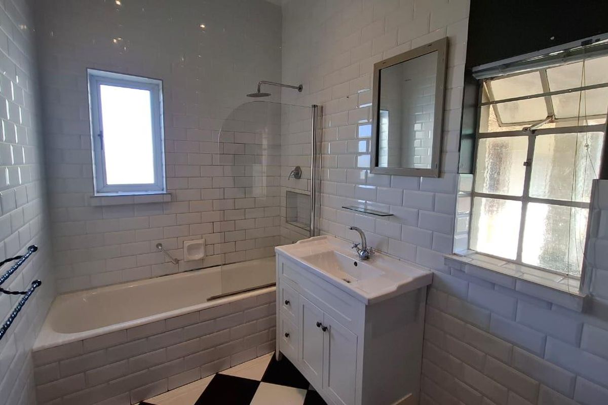 A bright bathroom featuring white subway wall tiles, a classic vanity unit, a bathtub with a glass shower screen, and a checkered floor.