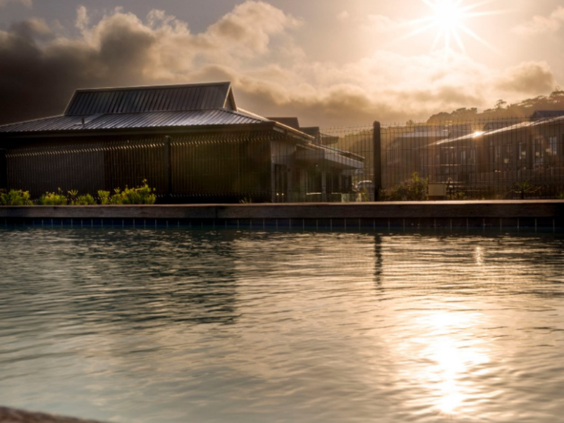 A serene view of the communal swimming pool area featuring timber decking and metal perimeter fencing at sunset.