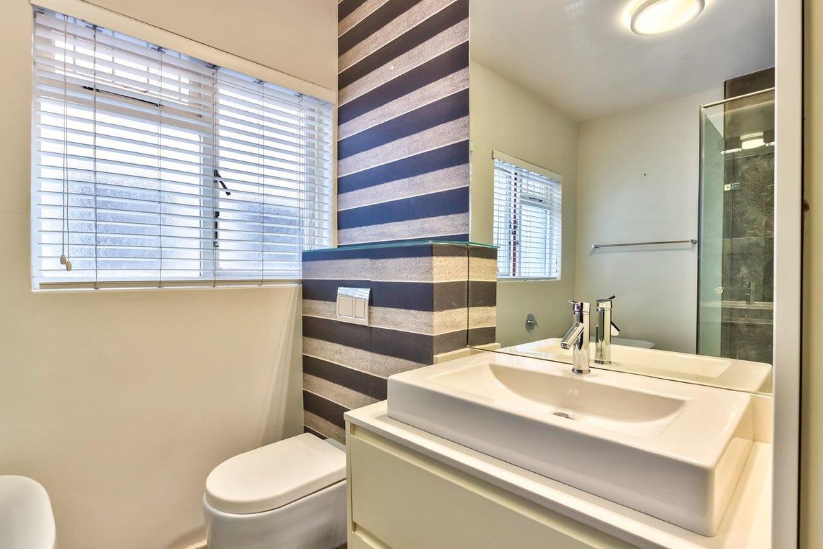 A clean, functional bathroom featuring a modern vanity, wall-mounted mirror, toilet, and a glass-enclosed shower with striped feature wall detailing.