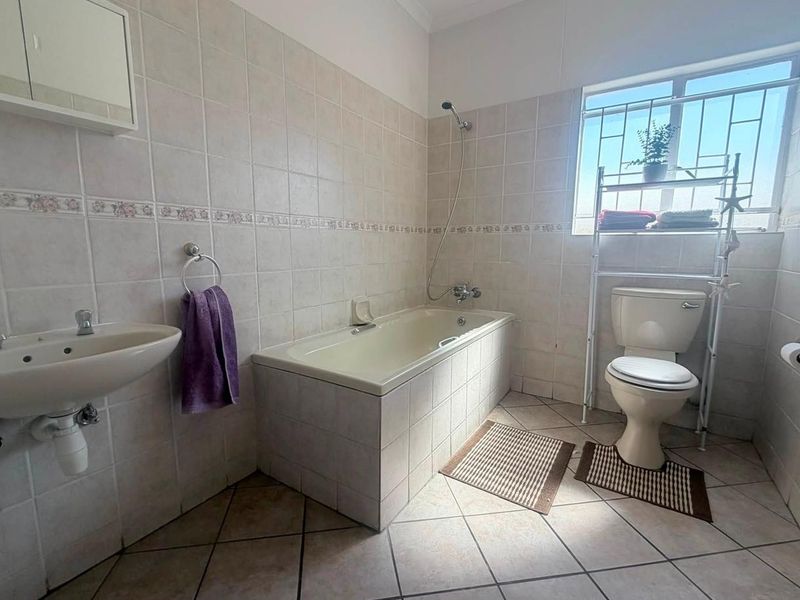 A functional bathroom featuring tiled walls and flooring, a standard bathtub with shower mixer, pedestal basin, and toilet, illuminated by natural light.