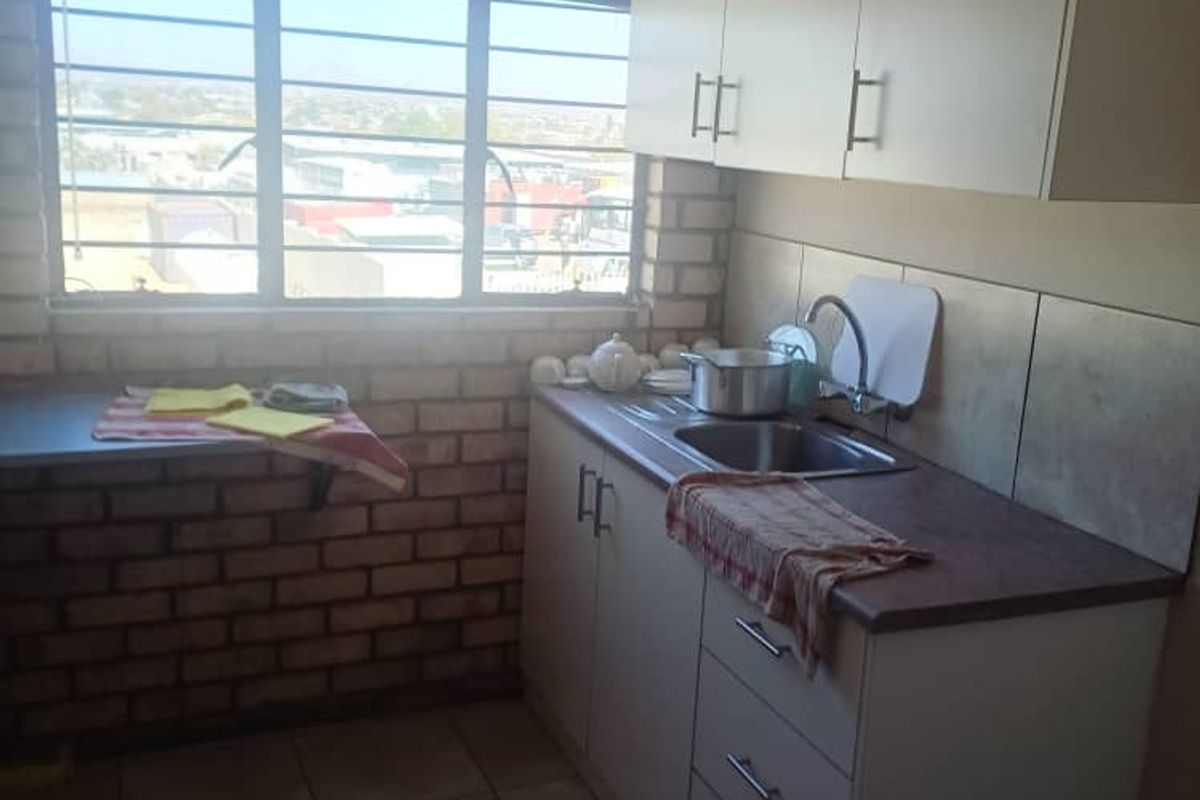 A compact kitchen area featuring built-in cabinetry, a stainless steel sink, tiled flooring, and an exposed brick wall with a window.