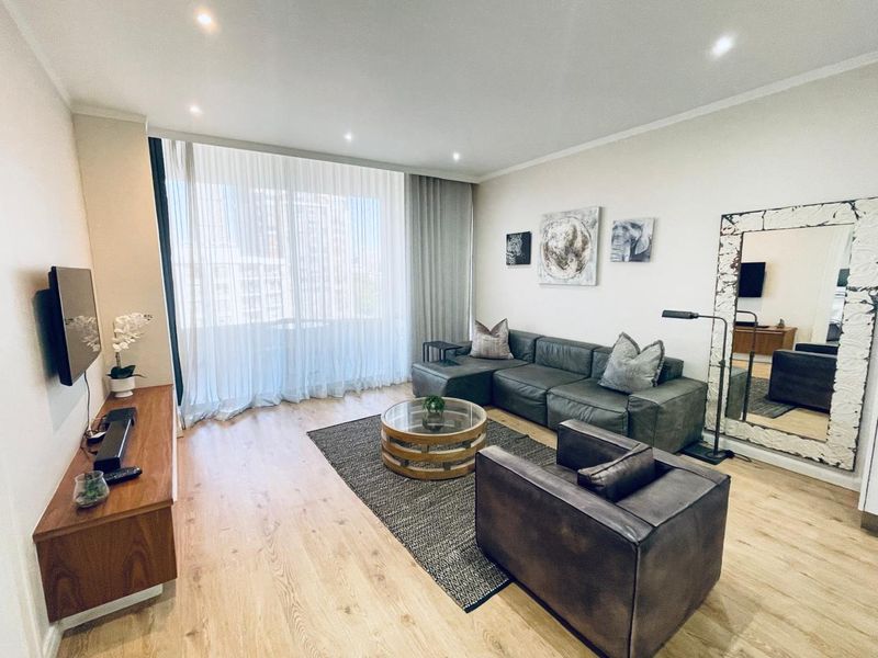 A well-maintained living room featuring modern laminate flooring, recessed downlighting, and large floor-to-ceiling curtains providing ample natural light.