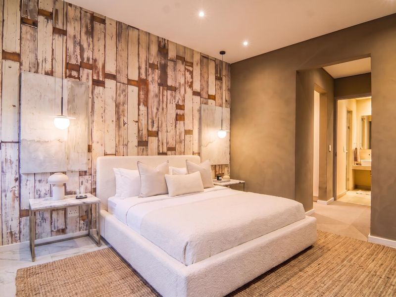 A modern, well-appointed bedroom featuring a textured feature wall, recessed downlighting, and elegant pendant fixtures, with an adjoining ensuite bathroom visible.