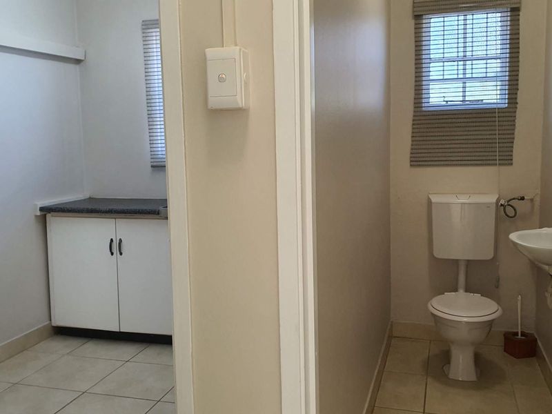 A functional office bathroom and adjacent utility area featuring tiled flooring, a standard toilet, wall-mounted basin, and built-in storage cabinetry.