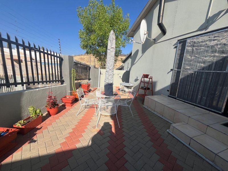 A paved outdoor patio area featuring metal security fencing, tiled steps leading to the entrance, and exterior wall-mounted infrastructure.