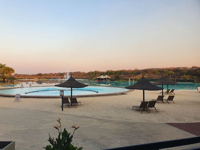 A large communal swimming pool area featuring a sandy beach, fountain, and paved patio space, offering a resort-style outdoor environment.