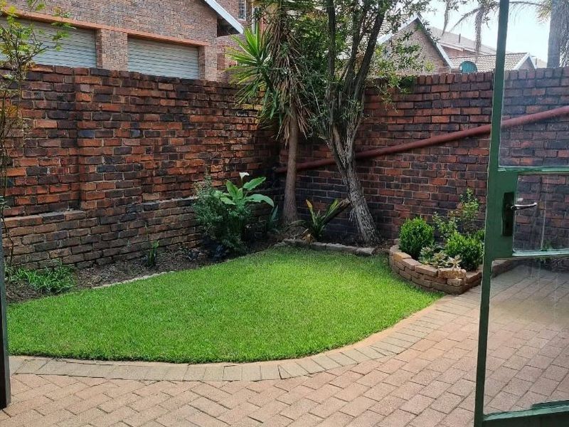 A private rear courtyard featuring a paved patio area, a small lawn, and a brick boundary wall, covered by a green corrugated roof structure.