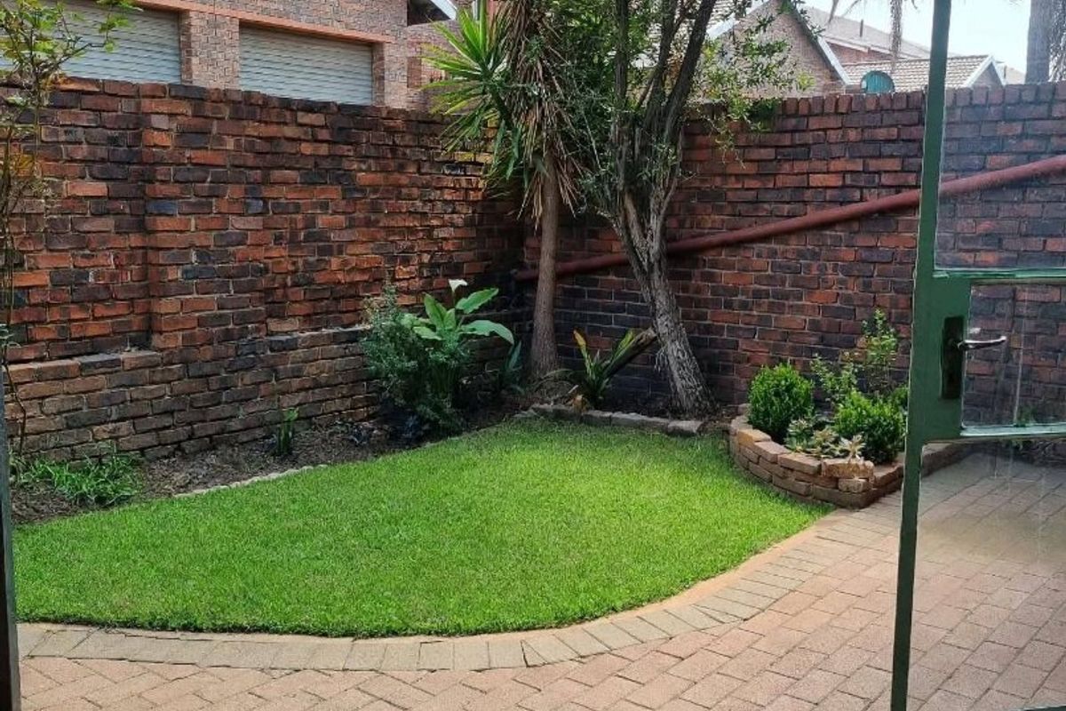 A private rear courtyard featuring a paved patio area, a small lawn, and a brick boundary wall, covered by a green corrugated roof structure.