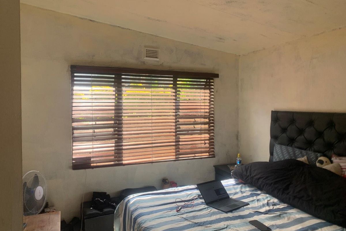A bedroom featuring a large window with fitted wooden blinds and a ventilation vent, requiring cosmetic updates to the walls and ceiling.