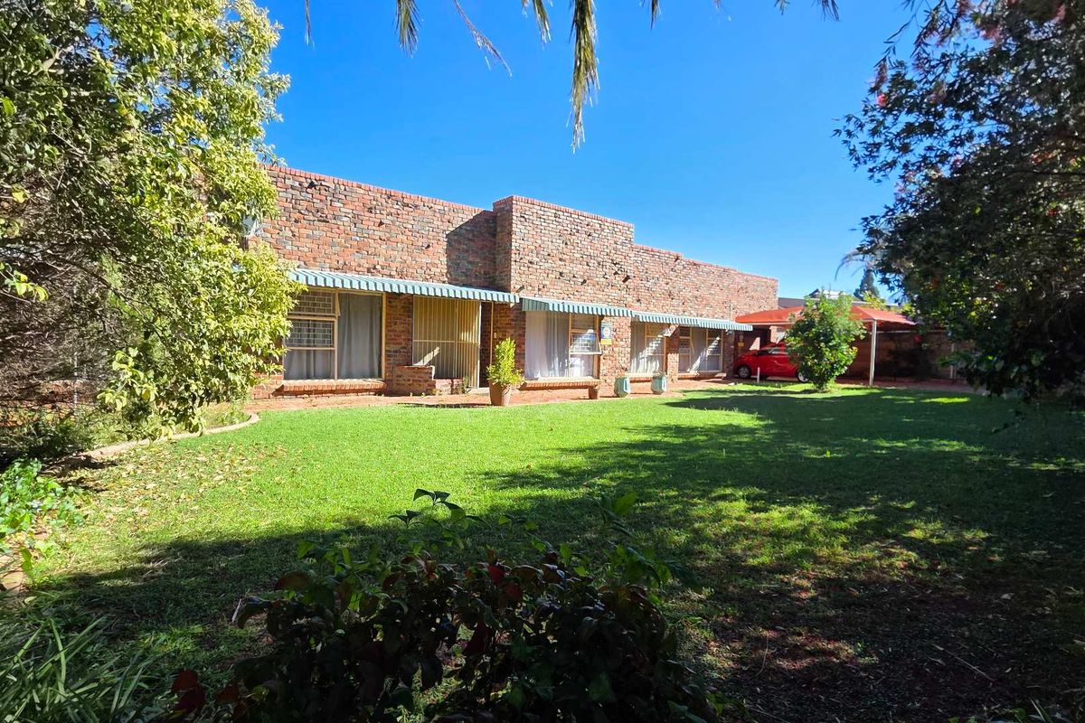 A spacious rear garden featuring a well-maintained lawn and a face brick house exterior with metal window awnings and an attached carport.