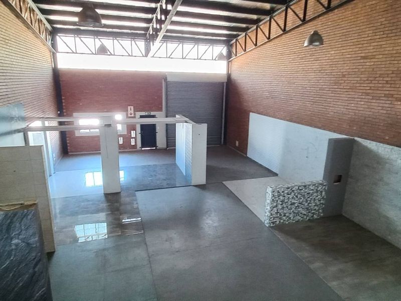 Spacious industrial warehouse interior featuring high exposed ceilings, brick walls, roller shutter door access, and partitioned floor areas suitable for various commercial or workshop uses.