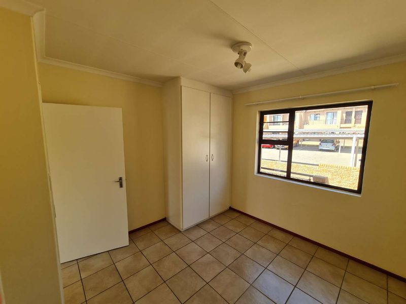 A clean, empty bedroom featuring neutral tiled flooring, a built-in wardrobe, and a large window providing natural light and views of the complex parking area.