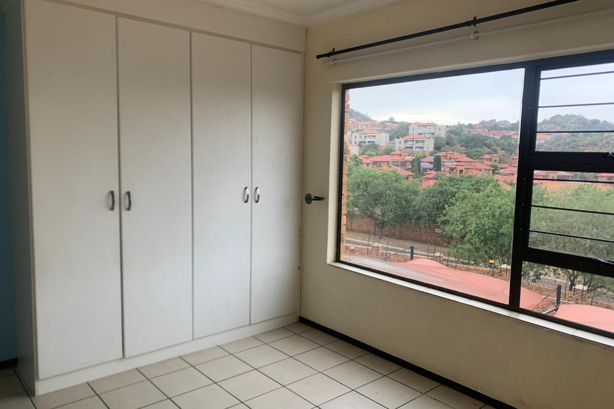 A bright, empty bedroom featuring built-in white cupboards, tiled flooring, and a large window offering an elevated view of the surrounding neighborhood.