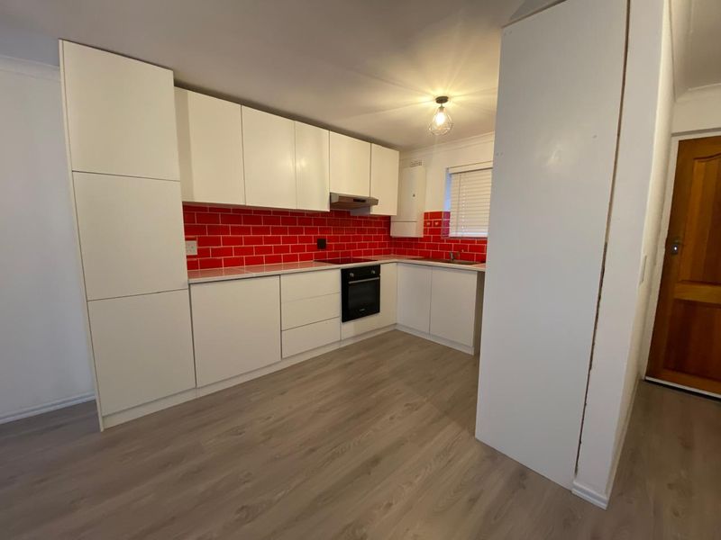 A clean, modern kitchen featuring white cabinetry, a bold red tiled splashback, integrated oven and hob, and light-toned laminate flooring.