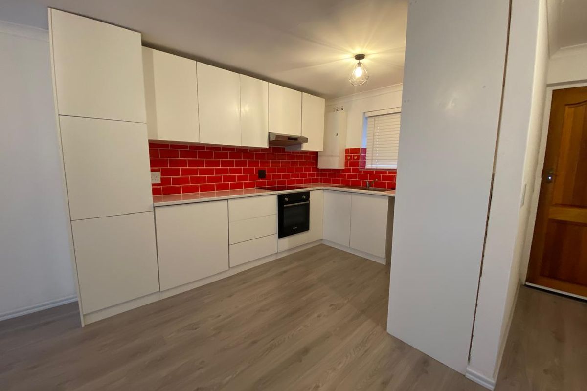 A clean, modern kitchen featuring white cabinetry, a bold red tiled splashback, integrated oven and hob, and light-toned laminate flooring.