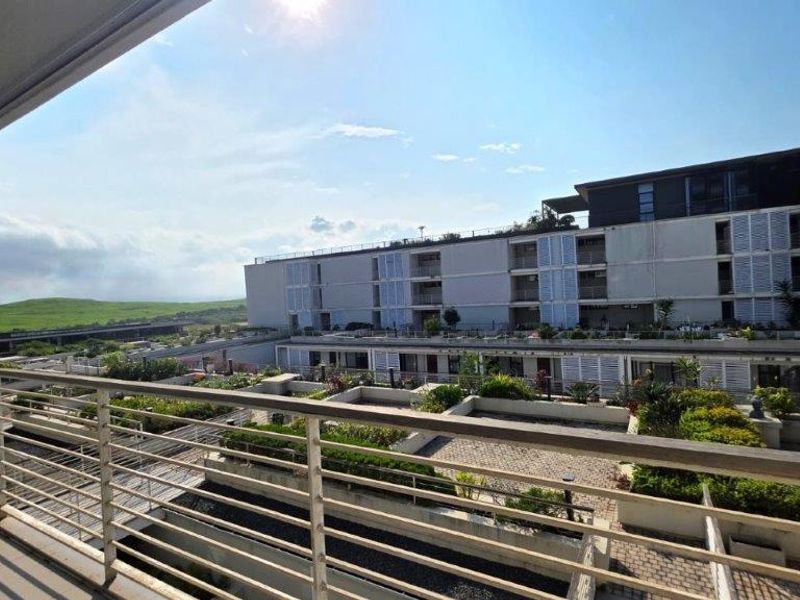A spacious balcony featuring a metal balustrade and sliding shutter screens, overlooking a landscaped terrace and surrounding apartment complex.