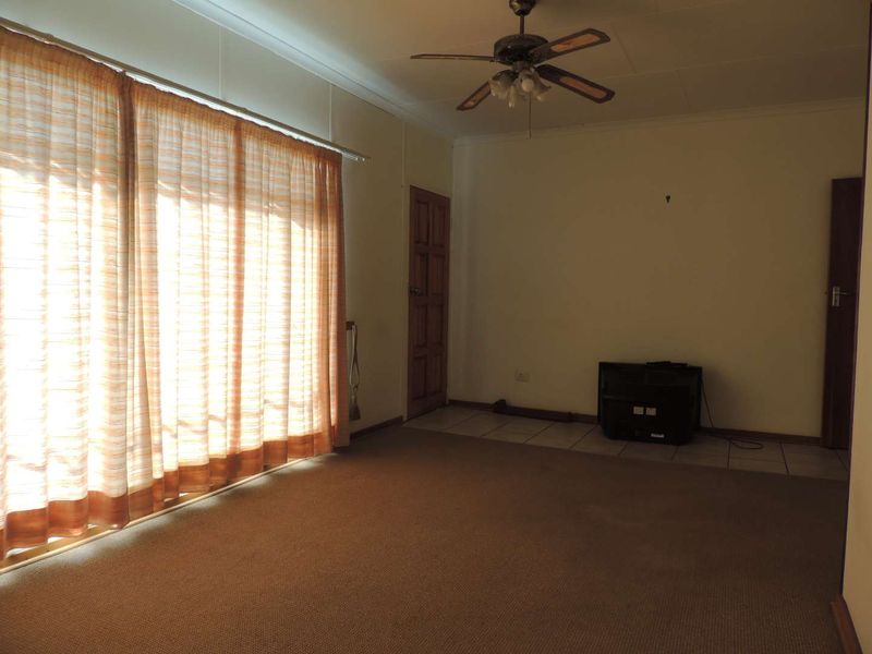 A spacious, empty living room featuring a ceiling fan, a mix of carpet and tile flooring, and large windows with curtains.
