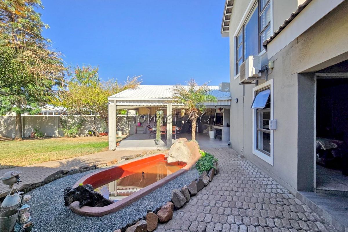 Rear garden view featuring a paved walkway, a decorative pond, and a covered patio area with a built-in braai.