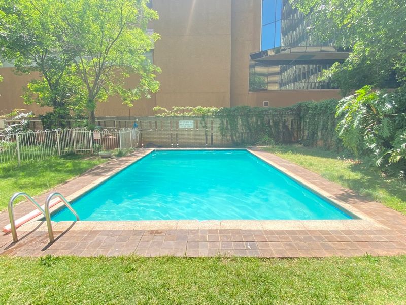 Outdoor swimming pool area featuring a rectangular pool with a ladder, surrounded by brick paving and a perimeter fence.