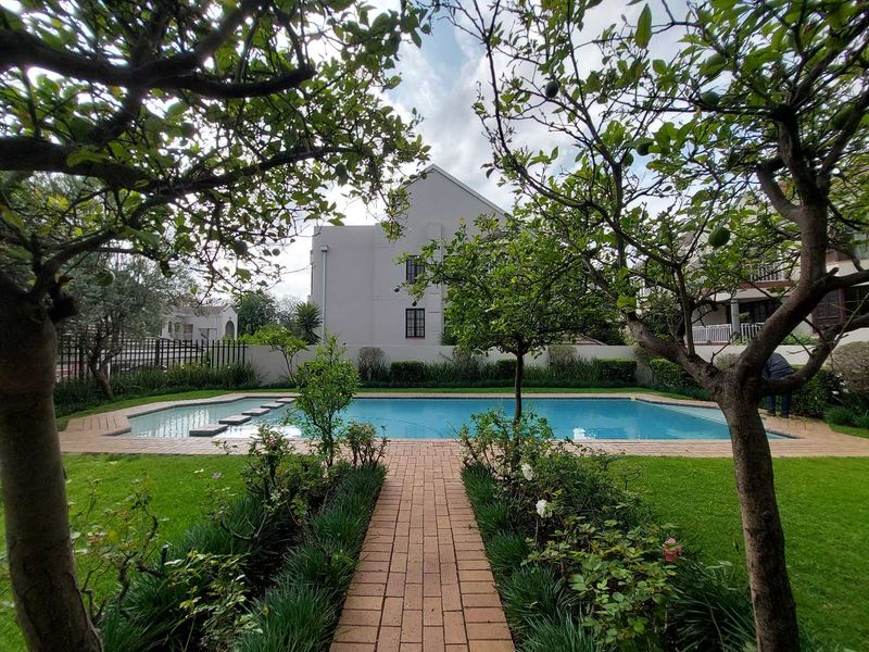 A well-maintained communal swimming pool area featuring a brick paved pathway, lush green lawn, and established trees within a secure complex setting.