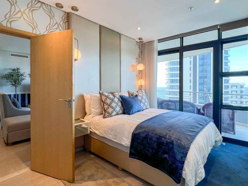 A well-appointed bedroom featuring a custom upholstered headboard wall, recessed lighting, and floor-to-ceiling glass doors leading to a private balcony with coastal views.