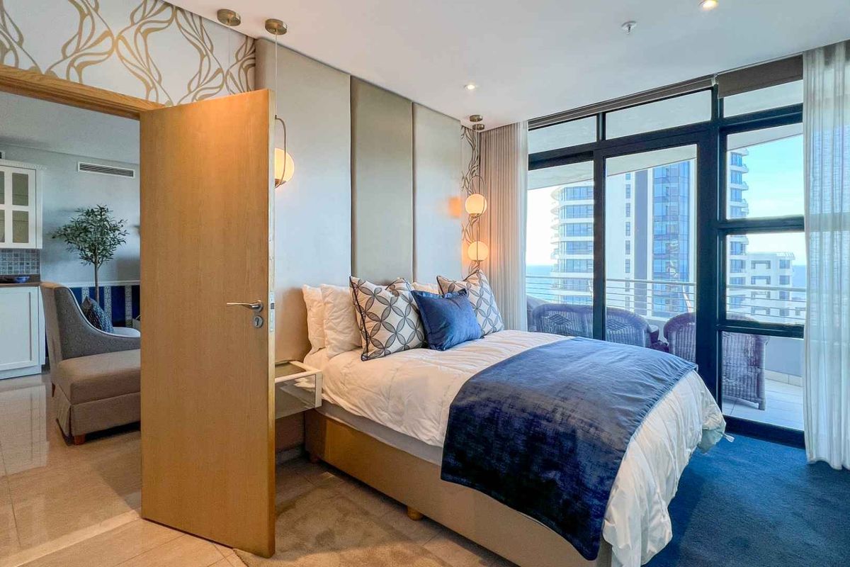 A well-appointed bedroom featuring a custom upholstered headboard wall, recessed lighting, and floor-to-ceiling glass doors leading to a private balcony with coastal views.