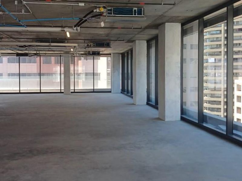 An open-plan office space featuring an industrial aesthetic with exposed concrete ceilings, ducting, and expansive floor-to-ceiling windows providing ample natural light.