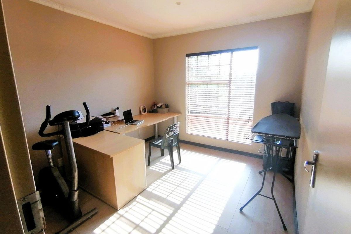 A functional bedroom or home office space featuring light-toned flooring, neutral wall paint, and a large window fitted with horizontal blinds.